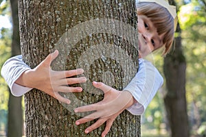 Portrait of a pretty smiling child girl in a paper crown on her head embracing a tree trunk in summer park. Love and care about