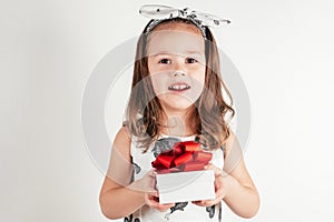 Portrait of smiling little girl in white dress with bow on head hold white gift box with red ribbon on white background