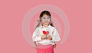 Portrait of pretty smiling preteen girl holding paper heart, happy looking at camera, posing isolated over pink