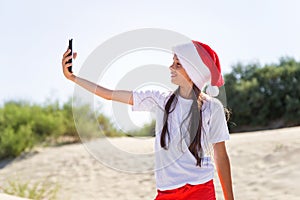 Portrait of a pretty smiling teenage girl in a red Santa hat and taking selfie on a sand tropical beach
