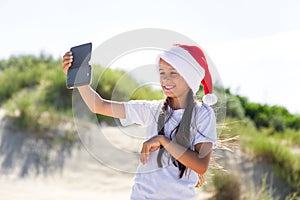 Portrait of a pretty smiling teenage girl in a red Santa hat and making selfie on a sand tropical beach