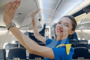 Portrait of pretty stewardess in blue uniform smiling at camera while closing hand luggage compartment, staying on the