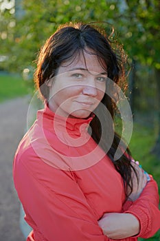 Portrait of a pretty woman 30-35 years old in a red ordinary windbreaker, in backlight, looks positively into the lens, brunette,