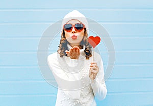 Portrait pretty woman blowing red lips sends air kiss holds lollipop heart wearing a heart shape sunglasses, knitted hat