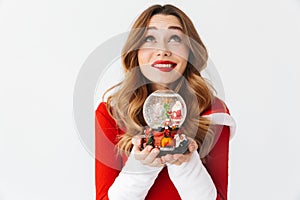 Portrait of pretty woman 20s wearing Santa Claus red costume smiling and holding Christmas snow ball, isolated over white