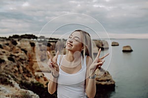 Portrait of pretty woman with victory gesture posing on rocky beach smiles to the camera.
