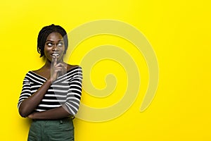 Portrait of a pretty young african woman showing silence gesture standing isolated over yellow background