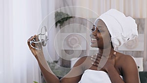 Portrait of pretty young african woman with white towel on head applied clay mask on face while watching in the mirror