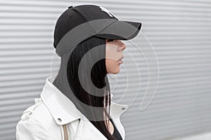 Portrait of pretty young beautiful woman in a stylish white leather jacket in a fashionable black baseball cap near a metal wall