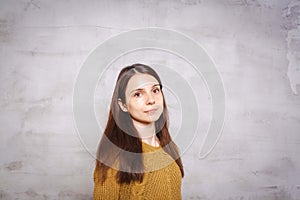 Portrait of pretty young brunette woman with long hair in amber pullover, stands in front of gray concrete background. Lifestyle