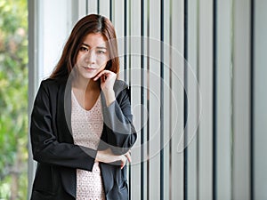 Portrait of pretty young business woman in casual black suit standing in the office with arm touch a chin, happy smiling and