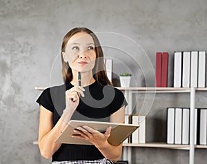 portrait of pretty young caucasian woman thinking with pen and tablet in hands, book shelf with copy space at background. pretty