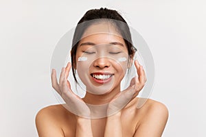 Portrait of pretty young chinese lady applying moisturizing cream on cheeks, posing over white background