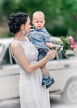 Portrait of pretty young female bride holding small baby boy with wedding roses bouquet at sunny summer park. Mother and her littl