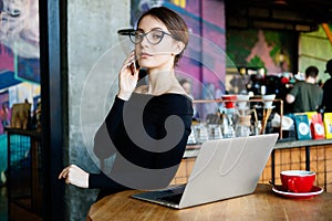 Portrait of pretty young freelancer woman on coffee shop. Beautiful lady with smart phone in her hands using laptop and looking at