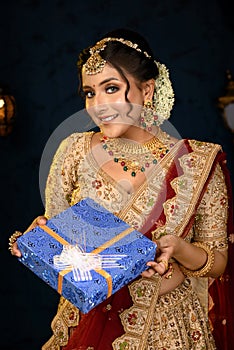 Portrait of a pretty young Indian woman dressed in traditional lehenga, gold jewellery and bangles holding gift box in hands on