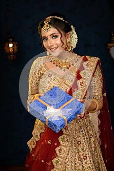 Portrait of a pretty young Indian woman dressed in traditional lehenga, gold jewellery and bangles holding gift box in hands on