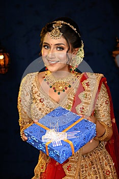 Portrait of a pretty young Indian woman dressed in traditional lehenga, gold jewellery and bangles holding gift box in hands on