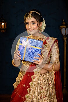 Portrait of a pretty young Indian woman dressed in traditional lehenga, gold jewellery and bangles holding gift box in hands on