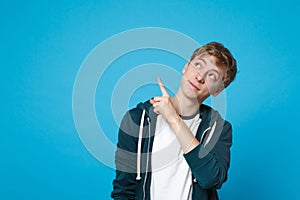 Portrait of pretty young man in casual clothes pointing index fingers aside and looking up isolated on blue wall