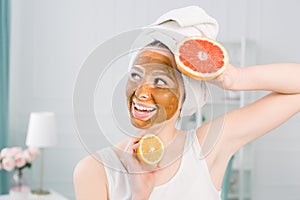Portrait of pretty young woman with clay mask on her face holding slices of lemon and grapefruit, facial care