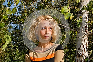 A portrait of a pretty young woman with a curly blond hair and blue eyes, standing near a birch in the park.