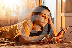 Portrait of pretty young woman is lying on bed and listening to music, putting headphones to her ear, looking into her
