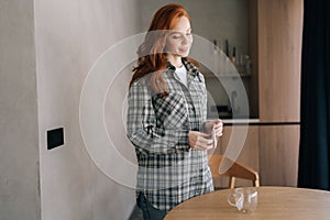 Portrait of pretty young woman opening coffee drip bag standing at kitchen table. Happy redhead female holding single