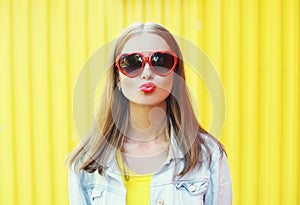 Portrait pretty young woman in red sunglasses blowing lips kiss over yellow