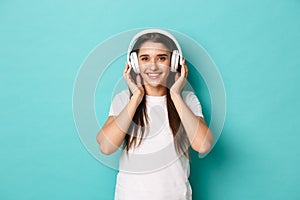 Portrait of pretty young woman in white t-shirt, smiling and listening to music in wireless headphones, standing over
