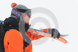Portrait of a professional athlete skier in an orange jacket wearing a black mask and with skis on his shoulder looks to