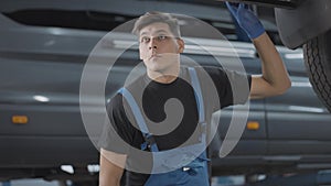 Portrait of professional auto mechanic checking fixing of vehicle standing under car in repair shop. Concentrated