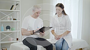 Portrait of professional female doctor consulting male patient in hospital ward. Ill mid-adult man signing documents for