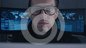 Portrait of professional hacker programmer in glasses at cyber security center filled with display screens.