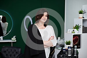Portrait of professional hairdresser in beauty salon. Young brunette female hair stylist standing in front of her workplace with