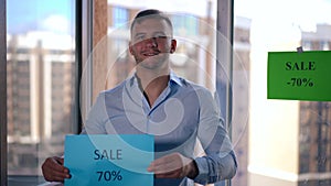 Portrait of professional real estate seller showing Sale banner smiling looking at camera. Confident young Caucasian man