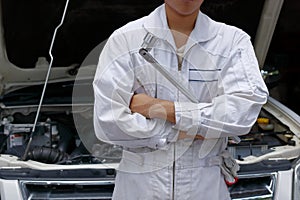 Portrait of professional young mechanic man in uniform holding wrench against car in open hood at the repair garage.