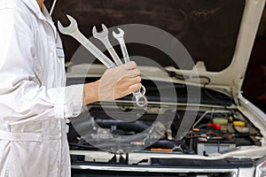 Portrait of professional young mechanic man in white uniform holding wrenches against car in open hood at the repair garage.