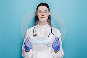 Portrait of professional young woman doctor or nurse in latex gloves holding medical protective face mask, looking at camera