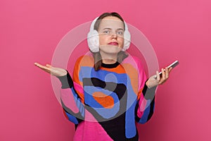 Portrait of puzzled confused uncertain woman wearing warm sweater and fur earmuffs standing isolated over pink background,