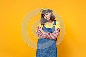 Portrait of puzzled disconcerted girl teenager in french beret, denim sundress holding hands crossed isolated on yellow