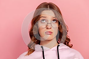 Portrait of puzzled female brunette teen in round eyeglasses looking up, thinking about information, doubting about choice