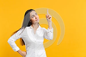 Portrait of puzzled pensive young woman in white casual shirt looking and pointing index finger up isolated on yellow