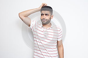 Portrait of query handsome bearded young man in striped t-shirt standing scratching his head, planing and thinking about something