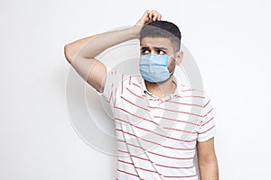Portrait of query young man with surgical medical mask in striped t-shirt standing scratching his head, planing and thinking about