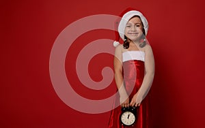 Portrait on red background of cute little girl, adorable child in Santa carnival outfit, holding alarm clock and smiling with