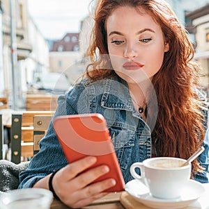 Portrait of red curled long hair caucasian teen girl sitting on a cozy cafe outdoor terrace on the street using the modern