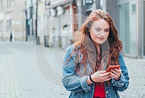 Portrait of red curled long hair caucasian teen girl walking on the street and browsing the internet using the modern smartphone.