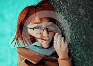 Portrait of a red-haired Asian woman in glasses and a warm beige coat leaning against a large cold stone rock on a winter day.
