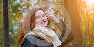 Portrait of a red-haired smiling girl in a jacket and scarf straightening her hair against the background of autumn nature and the